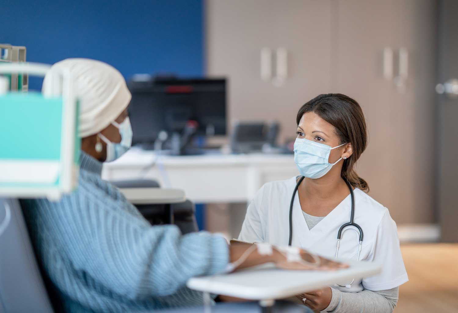A woman wearing a beanie and surgical mask sits on a chair as she receives chemotherapy. A nurse kneels as she explains a procedure.