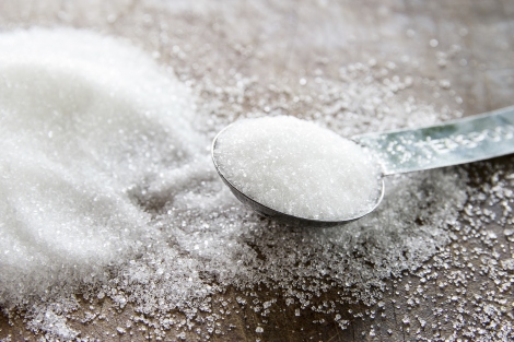 A teaspoon filled with sugar sits next to a pile of sugar