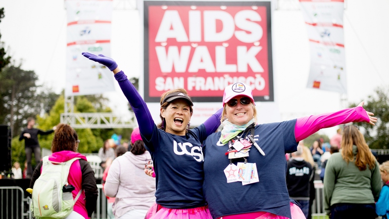 2 women wearing UCSF t-shirts and pink tutus pose in front of the AIDS Walk sign