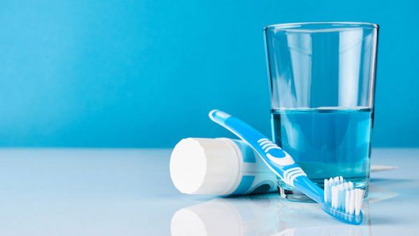 Photo of a toothbrush, tube of toothpaste, and a glass of blue mouthwash.