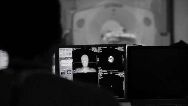 An MRI technician looks at brain scans in the foreground, while a subject is inside an MRI