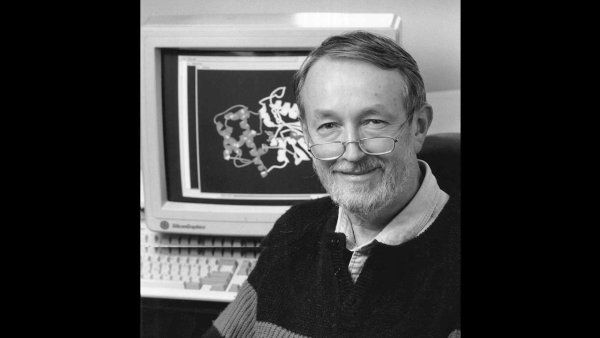 A greyscale photo of Henry Bourne. Behind him is a computer with a 3D molecular model on the screen.