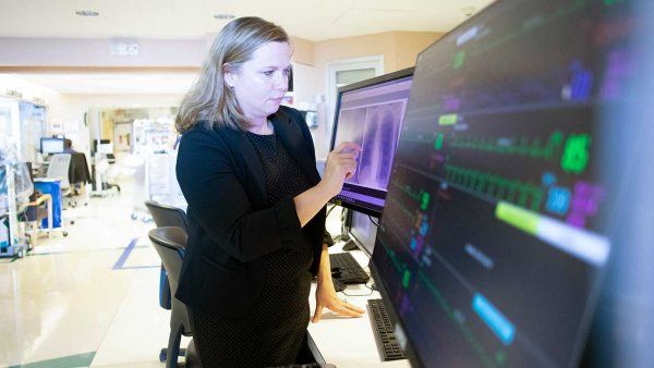 A doctor wearing a black dress and black blazer looks at an x-ray of a pair of lungs on a computer screen.