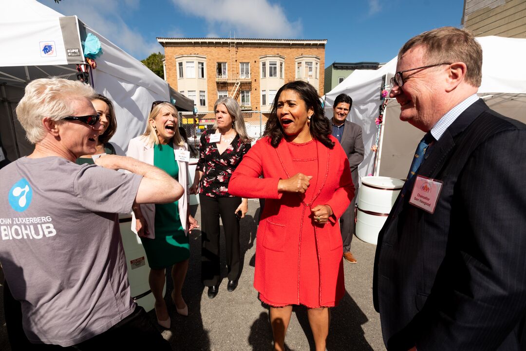 London Breed walks through a crowd while extending her elbow toward Joe DeRisi while the two smile at one another