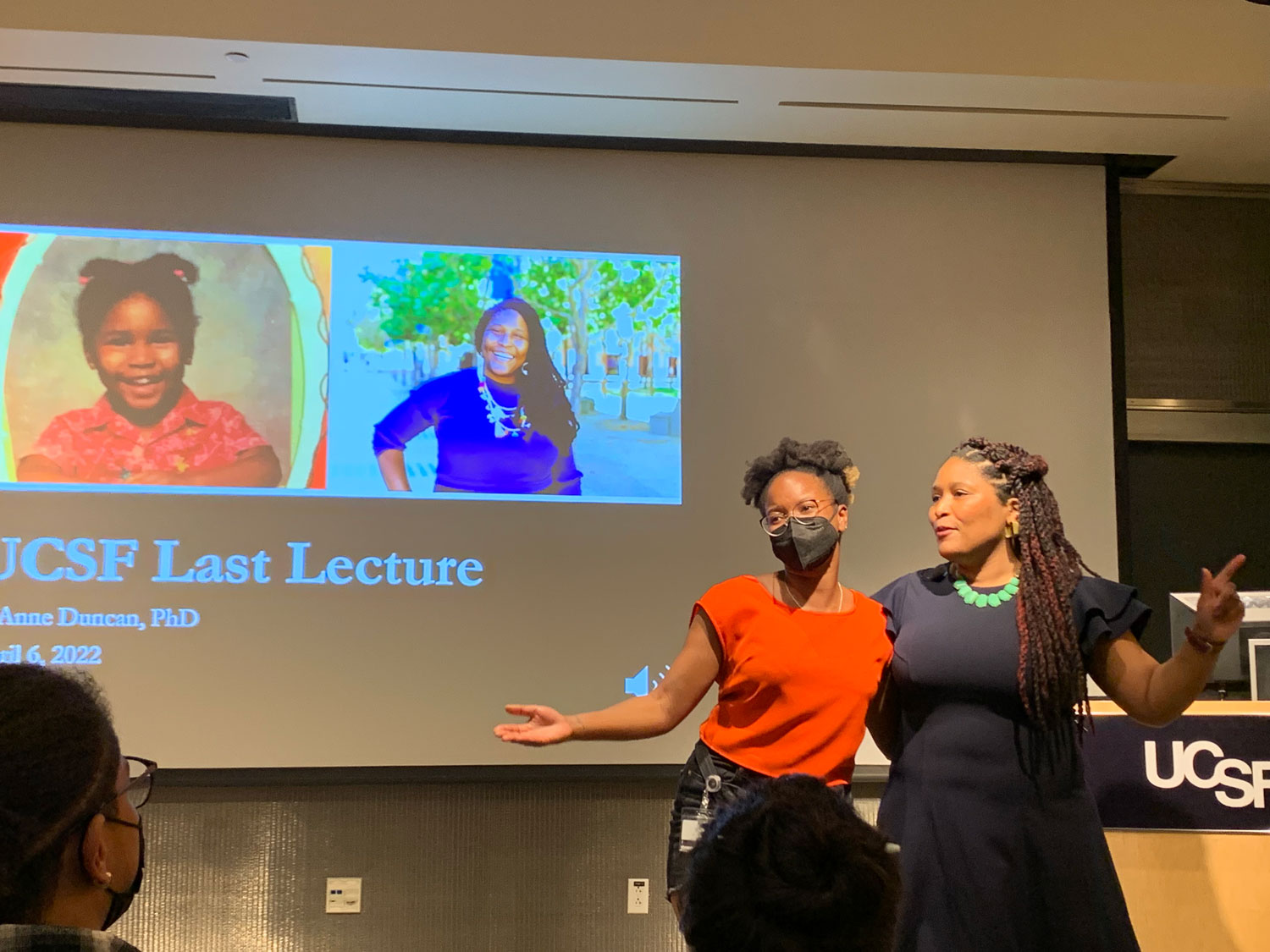 Dr. D'Anne Duncan stands on stage with an attendee of her 2022 Last Lecture event. In the background is a slide reading "UCSF Last Lecture", and a photo of Dr. Duncan as a child, and a current photo of her.