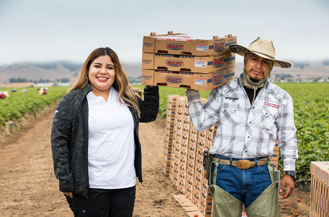 A nursing student on the left and an agricultural worker hold up boxes of fruit
