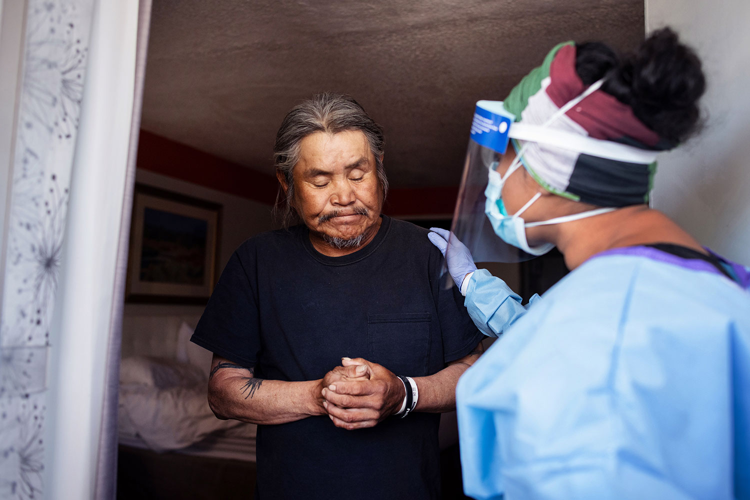 A health professional in a mask, face shield, and surgical gown places her hand on a man's shoulder. The man is a resident of the Navajo Nation