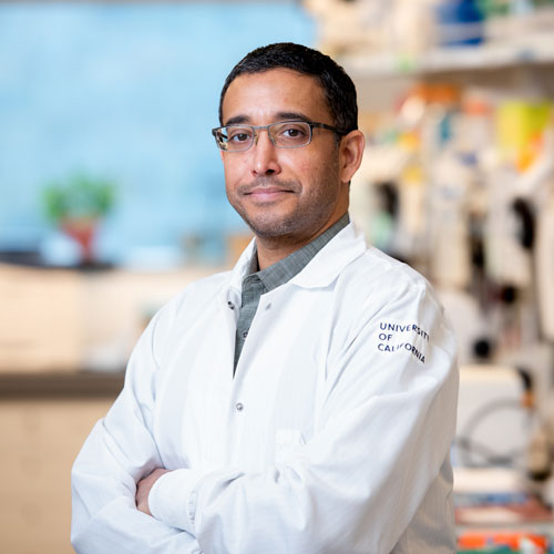 Devanand Manoli poses for a headshot in a lab, wearing a white lab coat. In the background are lab shelves full of equipment