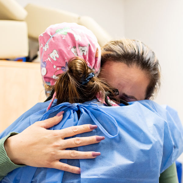 A dentist wearing a blue dental gown and a pink dental cap hugs the mother of a patient.