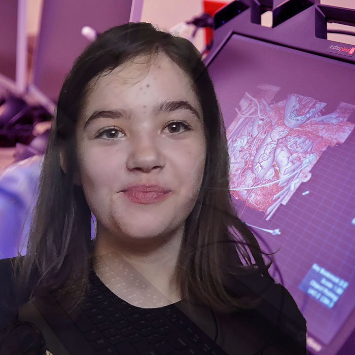 A young girl smiles. In the background is an image of a screen with a 3D computer heart model.