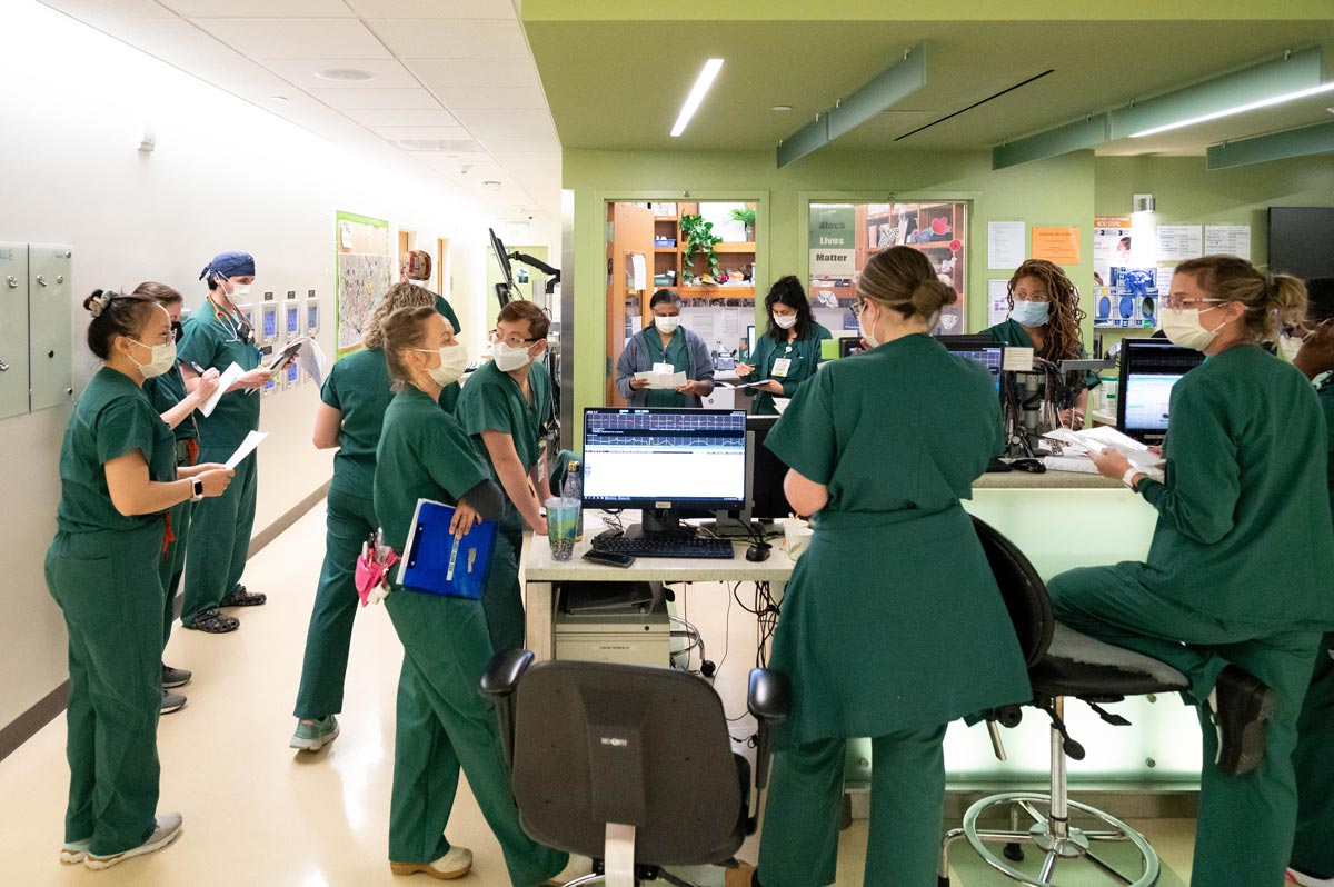 A team of medical staff wearing teal scrubs gathers on a hospital wing floor to disucss patients.