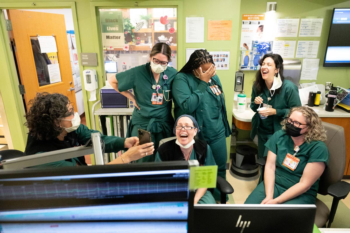 A group of medical staff wearing teal scrubs laugh together as one of them shows something on her phone.