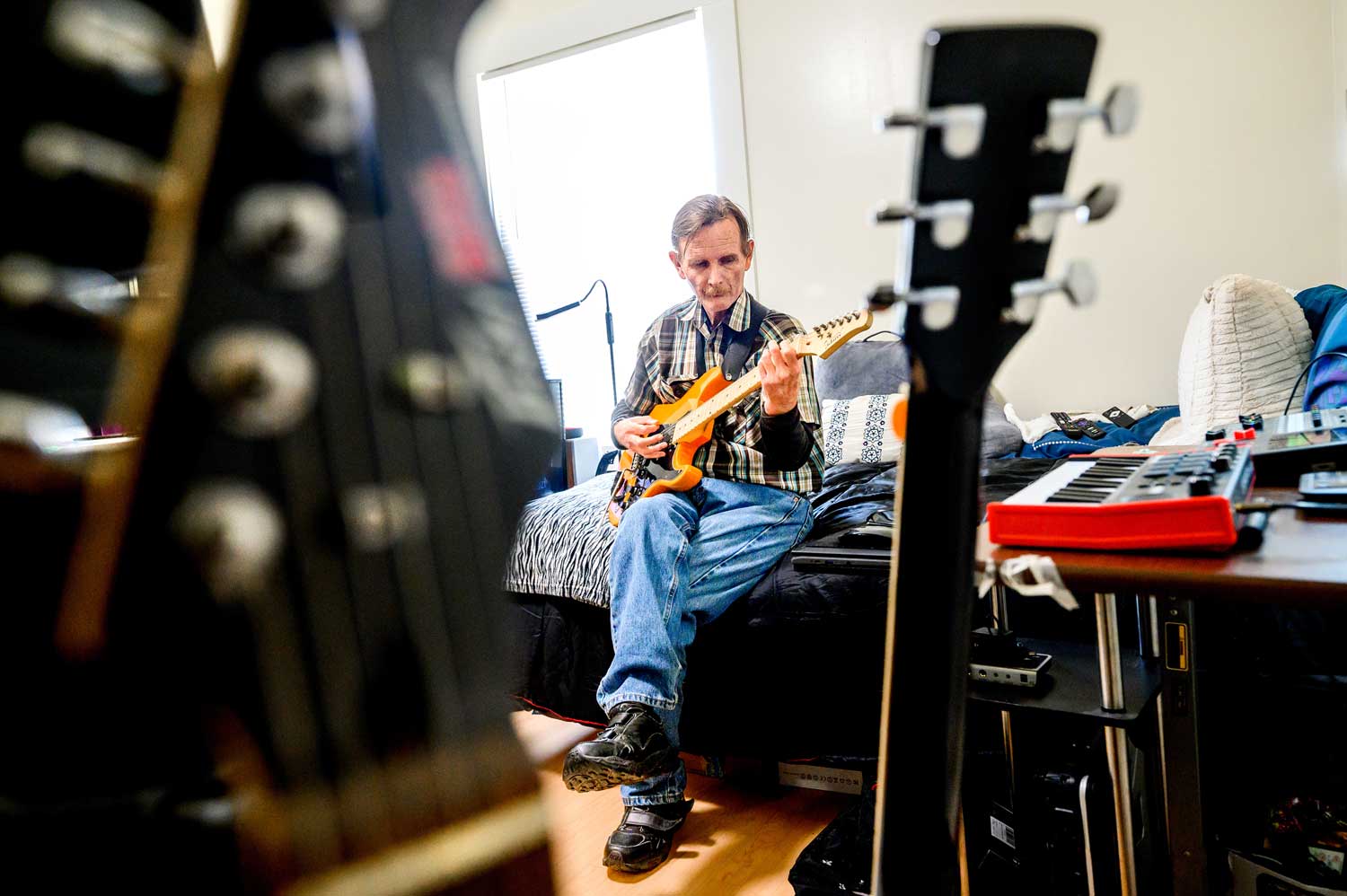 Bill sits on his bed and plays an orange electric guitar. In the foreground are the necks of two other guitars.
