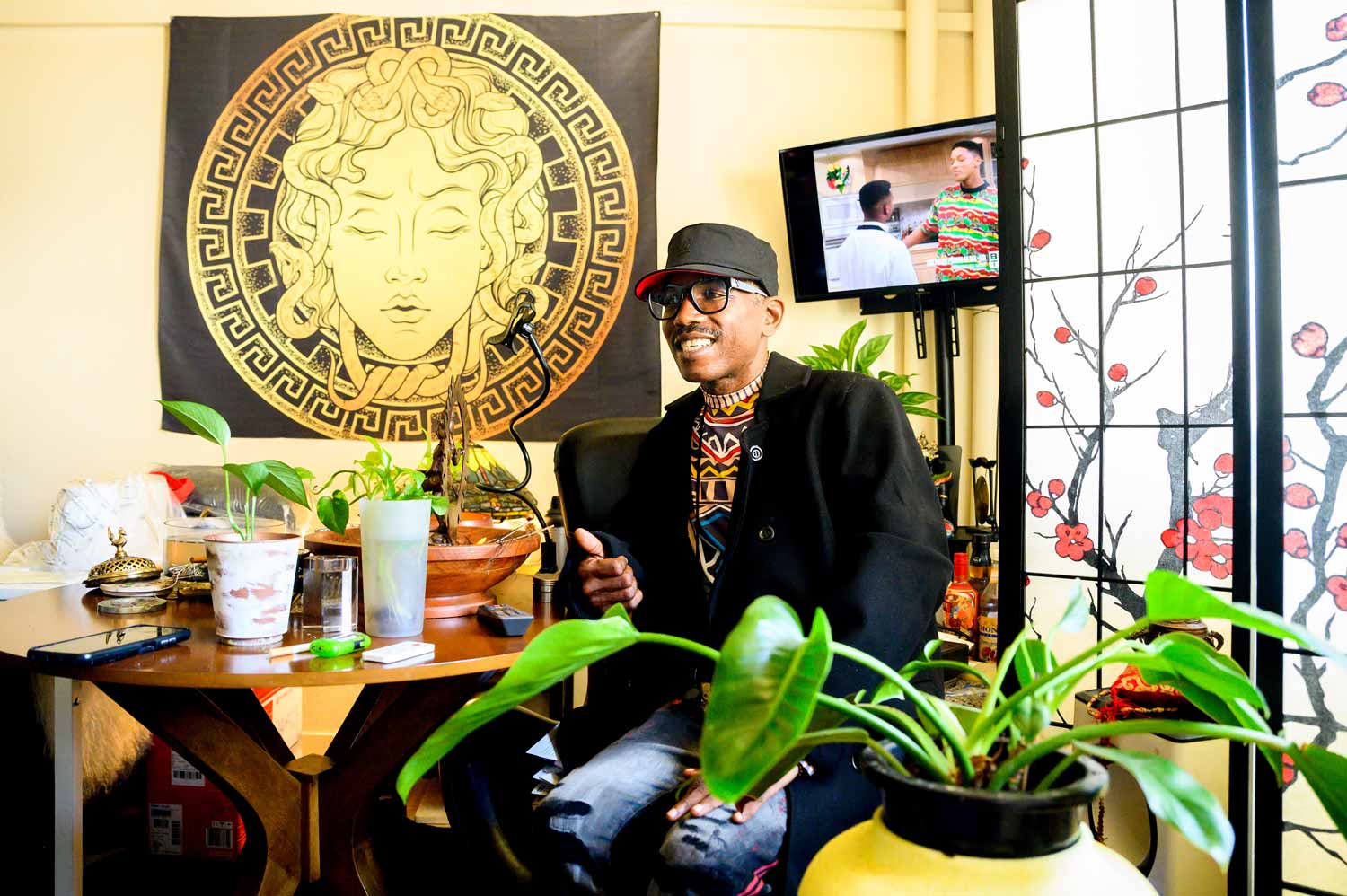 An African American man smiles as he sits inside his apartment. There are plant in the foreground, and a small television in the background.