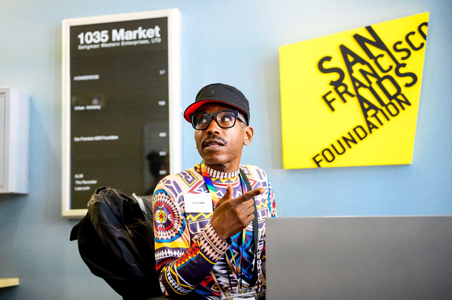 An African American man sits at a desk. In the background is a yellow sign that reads "San Francisco Aids Foundation."