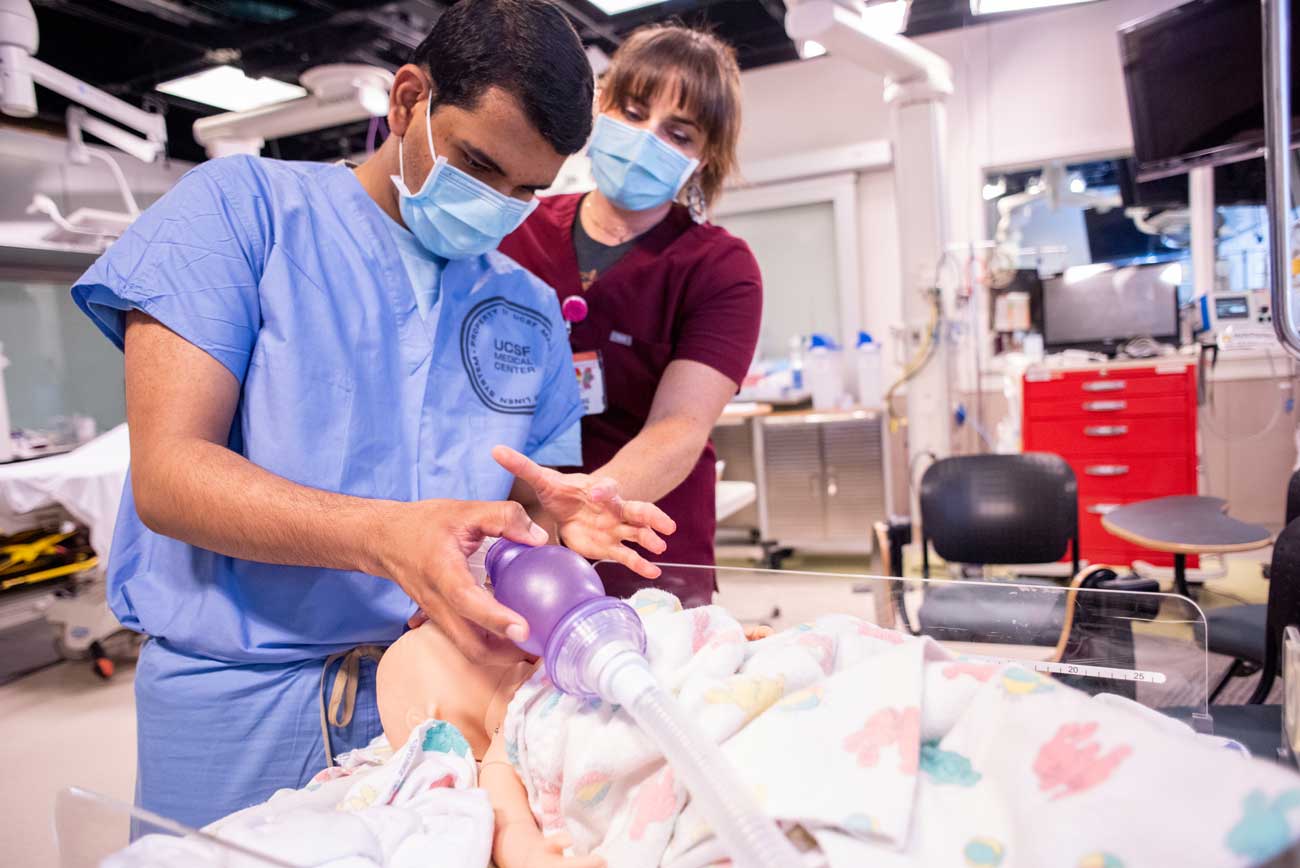 A medical student wearing blue scrubs practices delivering oxygen to a dummy while a doctor in red scrubs provides instruction.