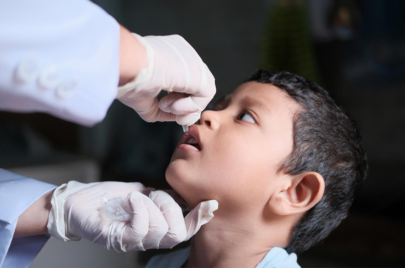 A pair of gloved hands deliver an oral polio vaccine with a dropper into a young boy's mouth.