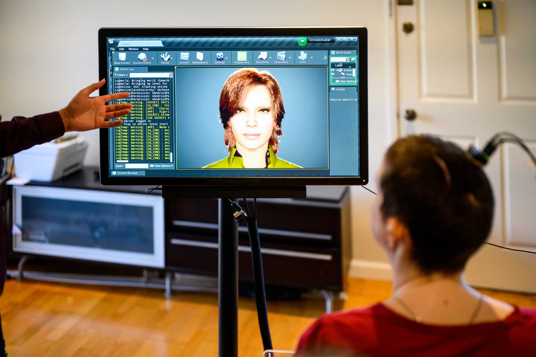A woman calmly looks at a screen with a 3D avatar that resembles her. The real woman has a buzzed haircut, and has wires protruding from her head that measure her brainwaves to produce speech from muscle movement.
