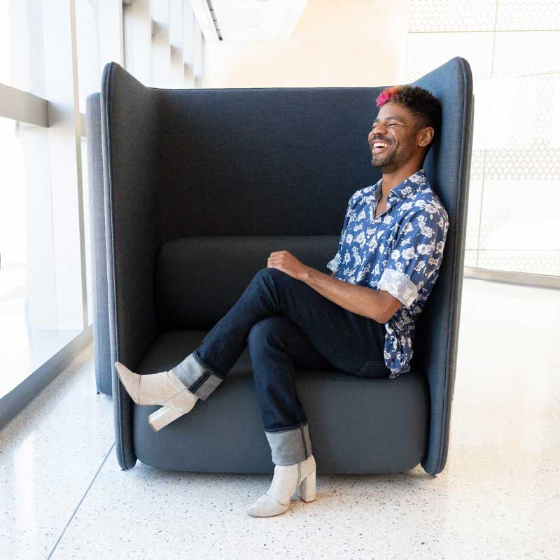 Chase Anderson smiles as he sits cross-legged on a fabric chair in a brightly-lit hallway.