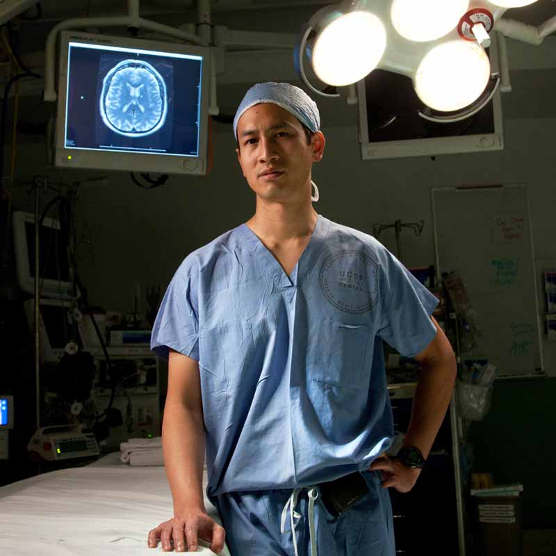 Eddie Chang, a neurosurgeon, wears a scrub uniform and a surgical cap while standing in an operating room. Behind him is a scan of a brain on a screen.