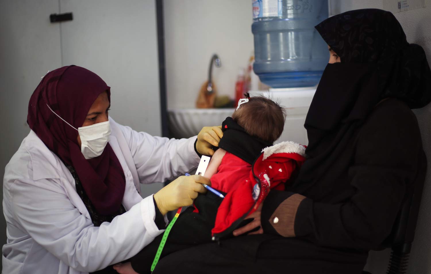 A doctor wearing a surgical mask examines a malnourished baby as her mother holds her.