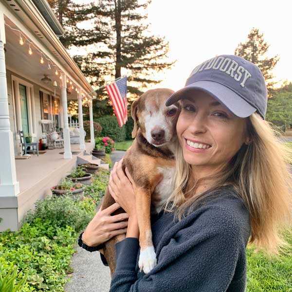 A woman wearing a baseball cap smiles as she holds her dog outside of a house.