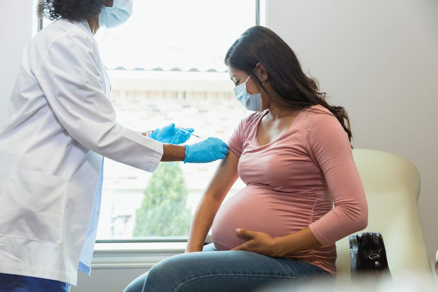 A doctor wearing a white coat and blue latex gloves injects a vaccine into a pregnant woman's arm.