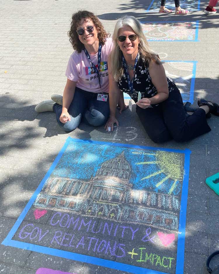 Two women who are UCSF employees sit on the ground next to their chalk drawing of a government building with the text "Commuity & Gov Relations + Impact."