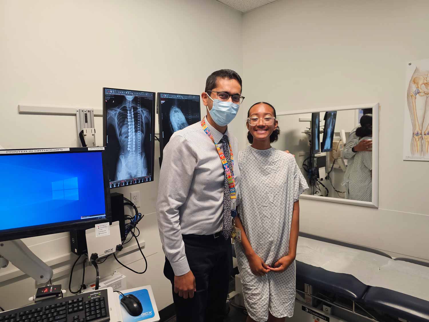 Dr. Ishaan Swarup poses for a photo with patient Malaya in an examination room. Behind them are computer screens showing x-rays of Malaya's spine.