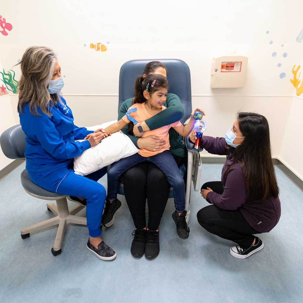 A mother and daughter sit together upright in an examination chair, while a nurse prepares the child for a blood draw and a medical professional provides a comforting distraction for the child.