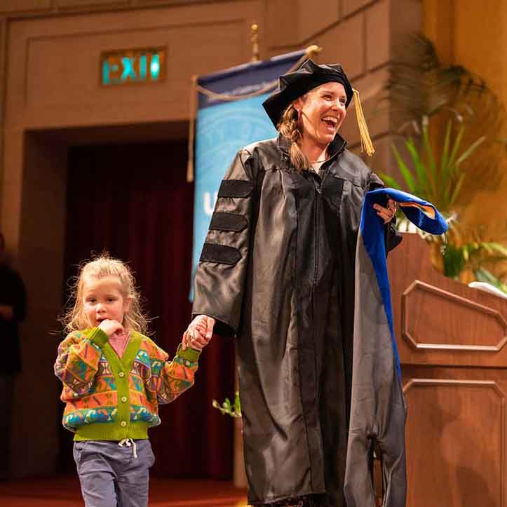A UCSF graduate division graduate in academic regalia walks across the stage holding her toddler daughter's hand as she receives her diploma.