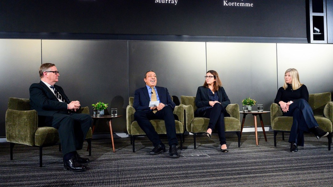 AI panelists sit under large screen on couches with the Chancellor