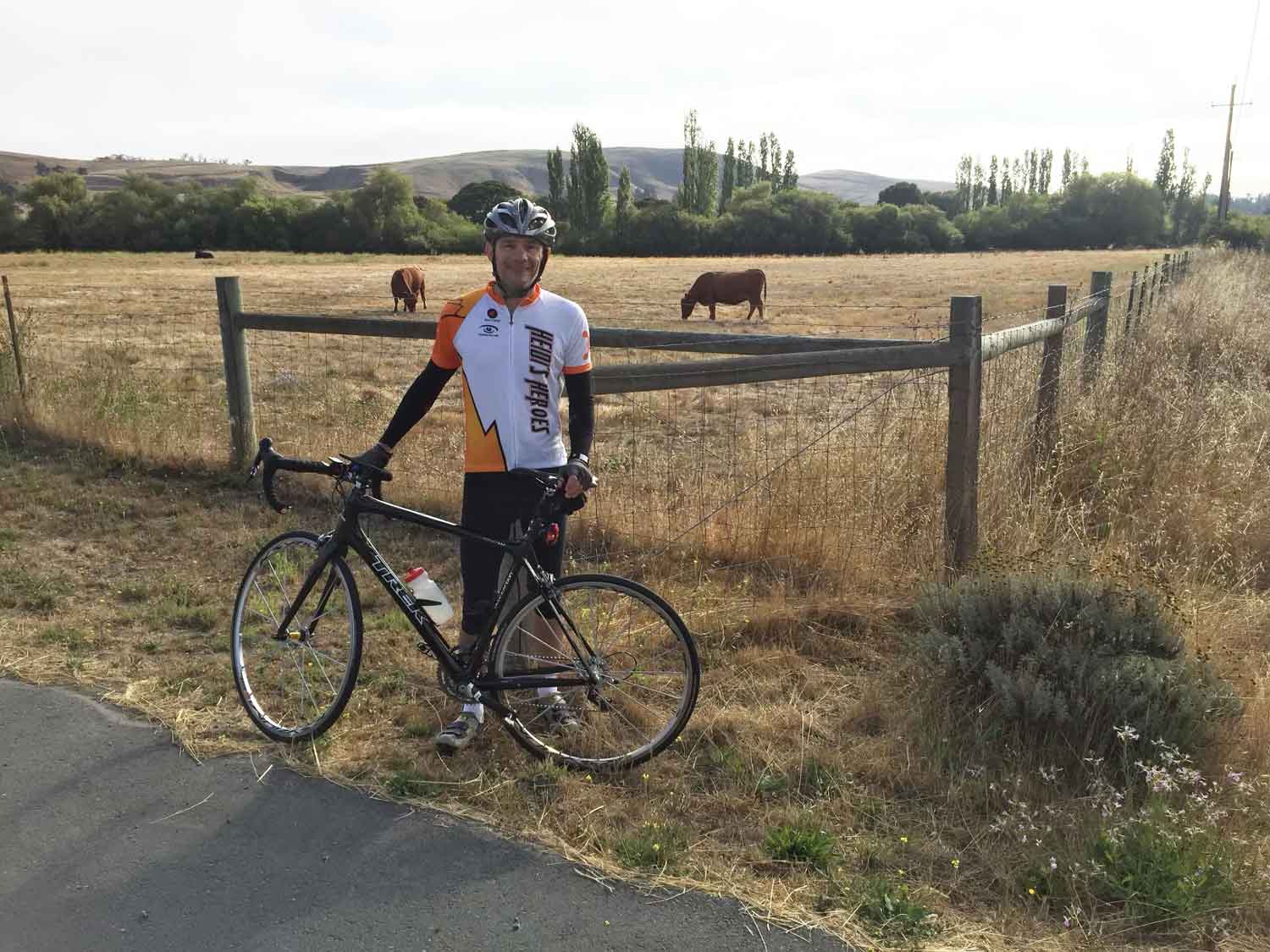 A middle-aged man stands with his bike in front of a pasture where a cow is grazing.