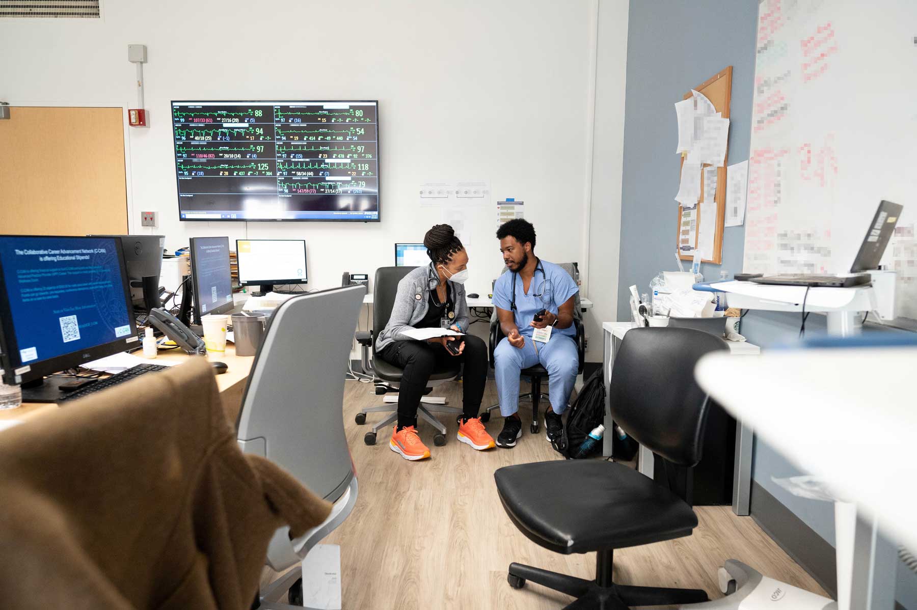 Two Black medical residents named Kelechi and Chris sit and discuss patients in a resident working area. They are surrounded by computers, chairs, and patient information on whiteboards.