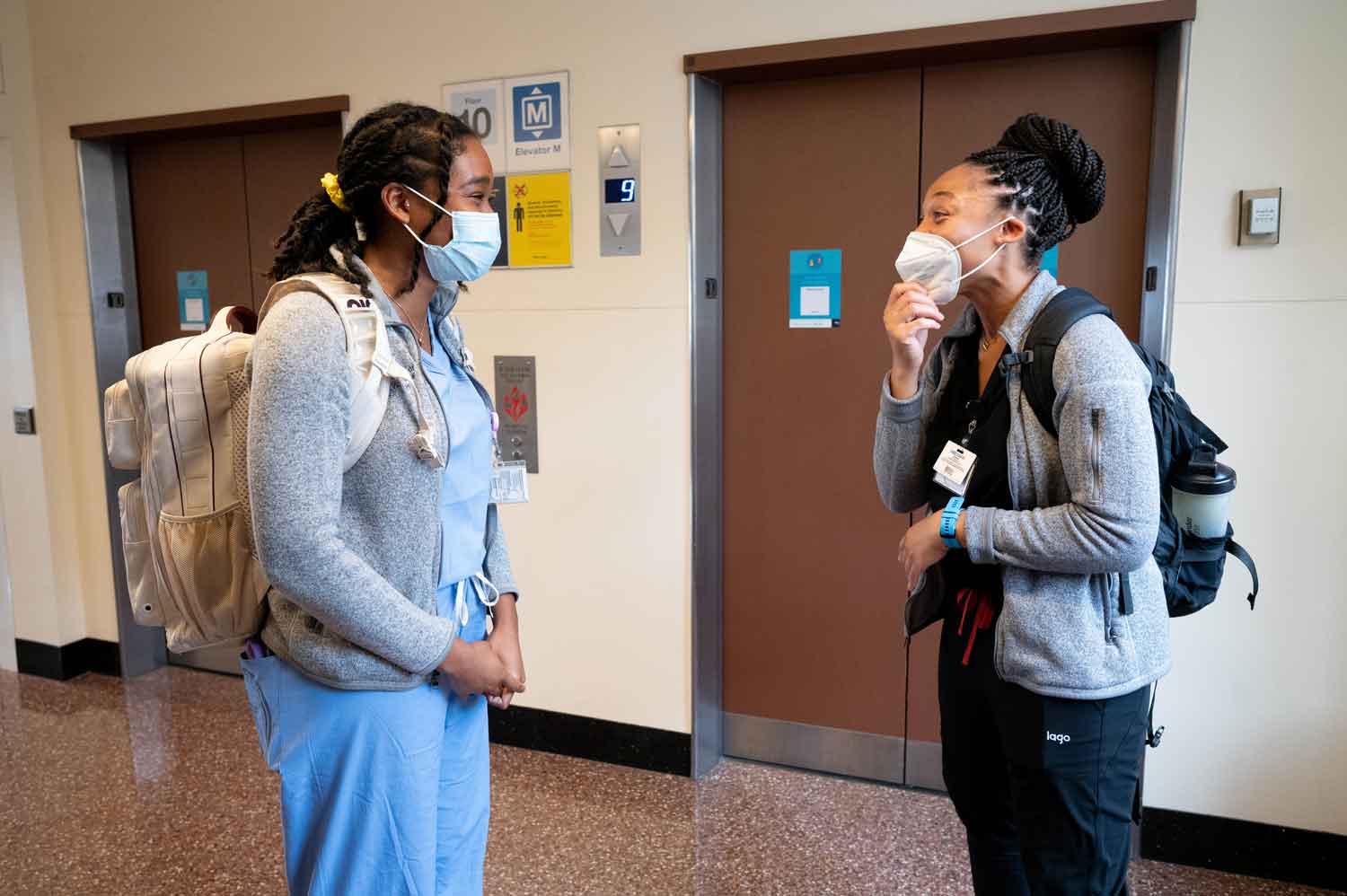 Two Black female medical residents named Jas (left) and Kelechi (right) smile as they greet each other in front of a set of elevators .They wear respiratory masks and medical scrubs.