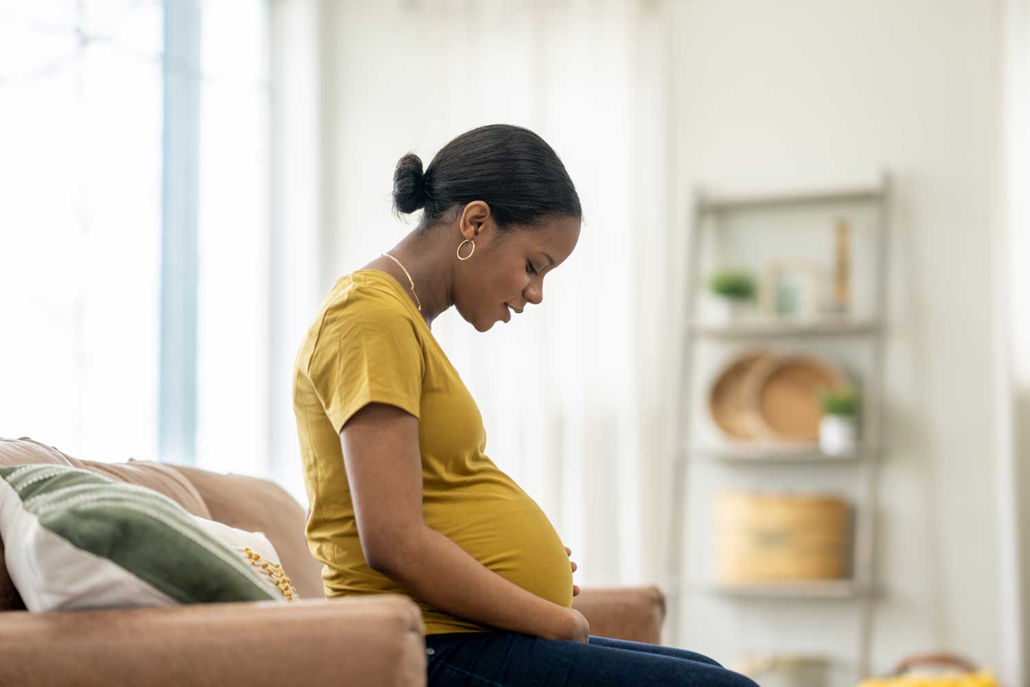 A Black pregnant woman sits comfortably on her couch at home and affectionately looks down at her growing stomach.