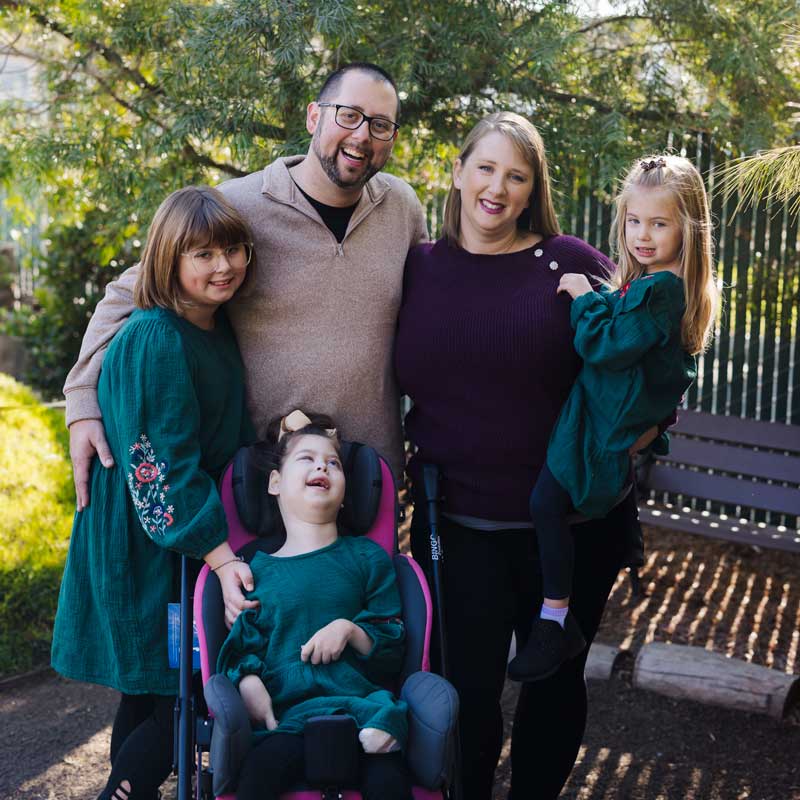 A family pose for a photo. They include a father, mother, and three daughters. One of the daughters, who has Gould Syndrome, smiles as she sits in her wheelchair.