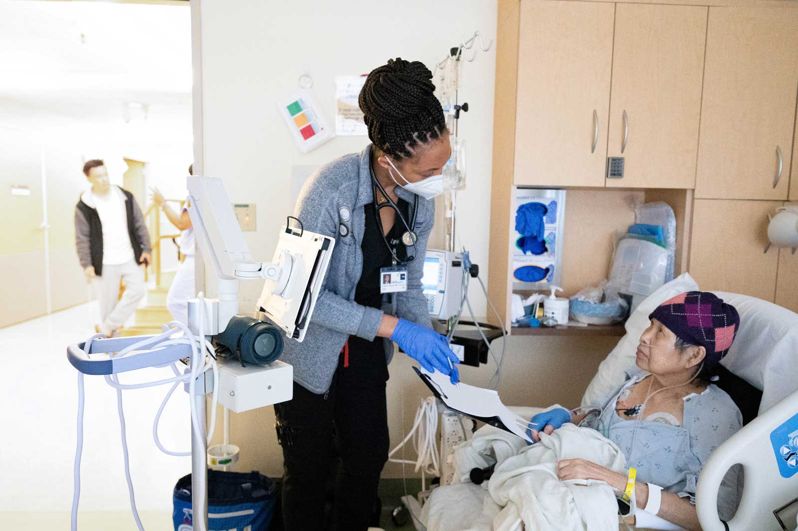 First-year resident Kelechi shows a patient paperwork on a clipboard. The patient is an elderly woman of Asian descent, and wears a beanie and hospital gown as she lies in hospital bed.