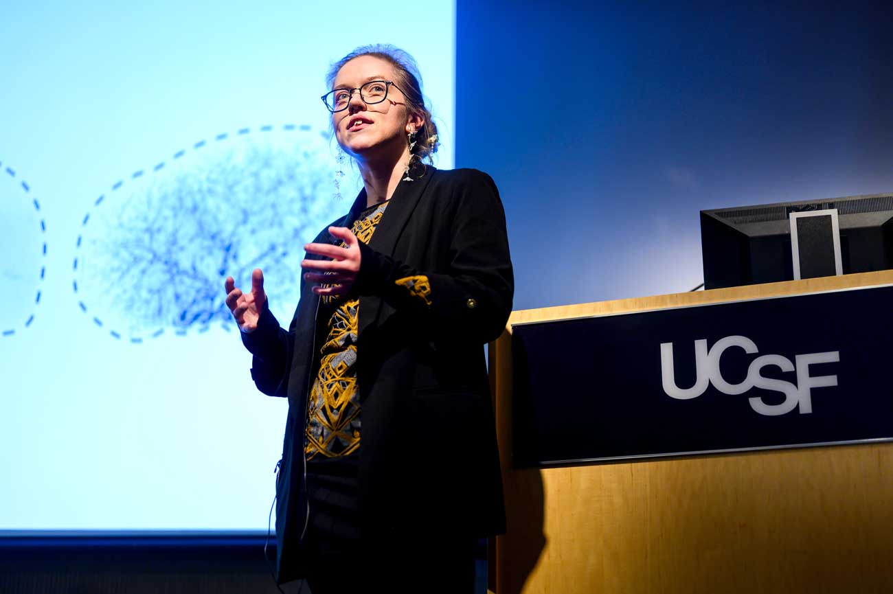 Simone Kurial presents her research onstage. Behind her is a projection screen with scientific images.