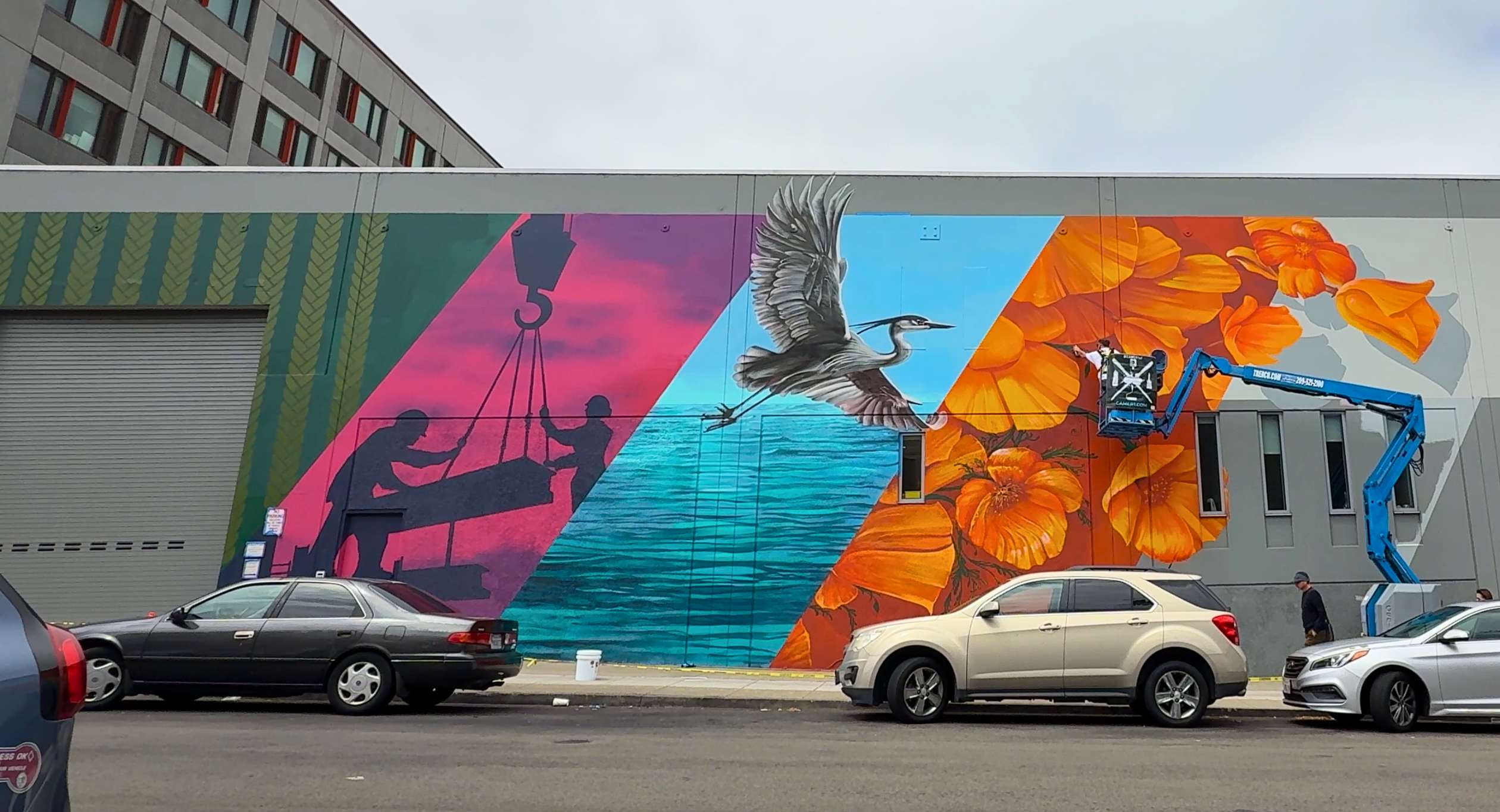 Cars parked in front of multi-panel mural on industrial building.