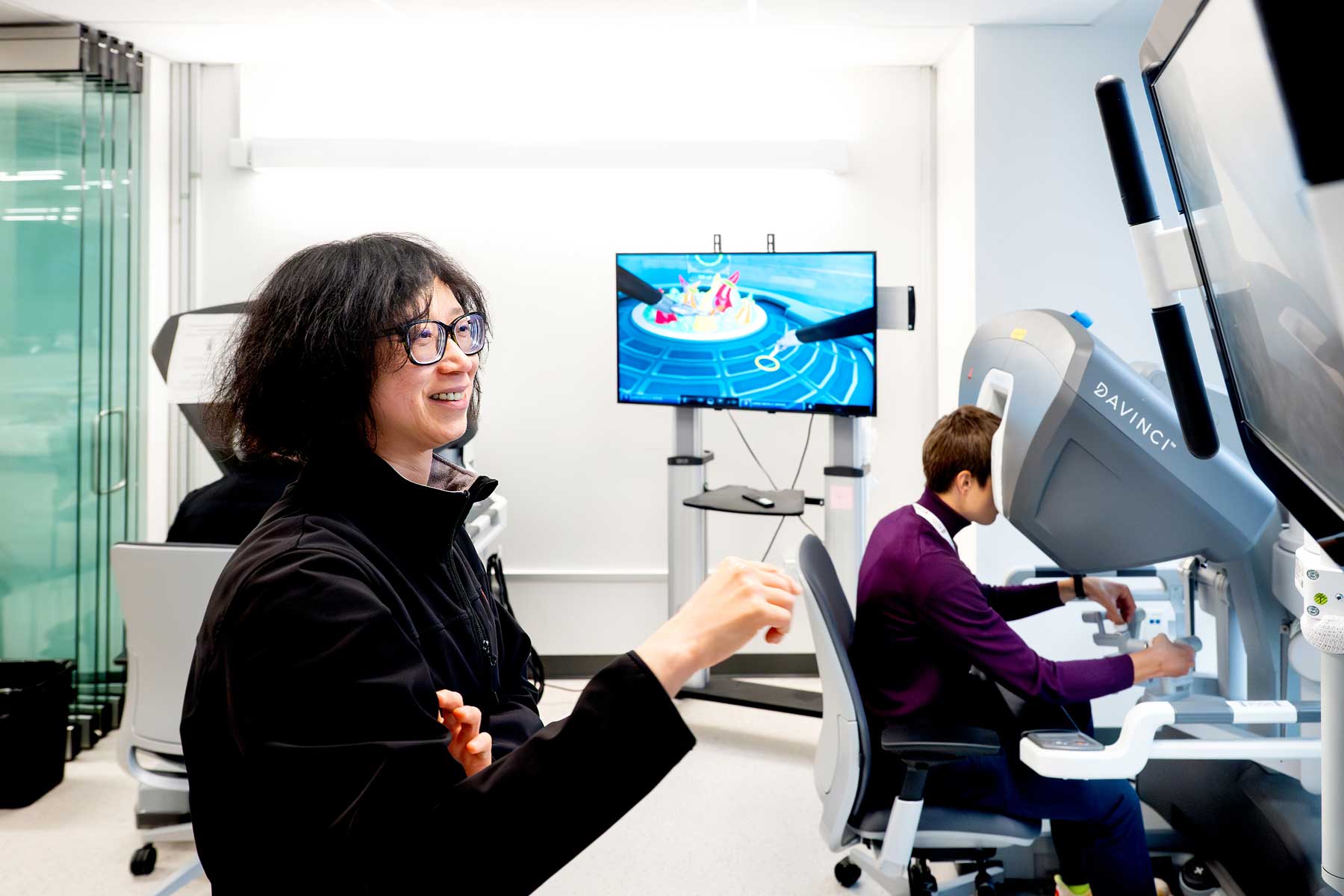 Dr. Hueylan Chern smiles while standing next to a surgical resident sitting at a Da Vinci surgical robot console. A monitor between them shows a close-up of a surgical tool.