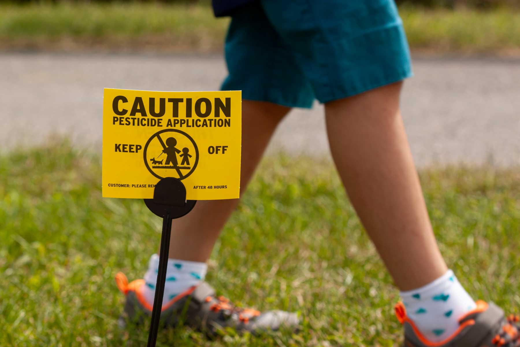 A child walks on grass with a warning sign that reads "Caution: Pesticide application, keep off."