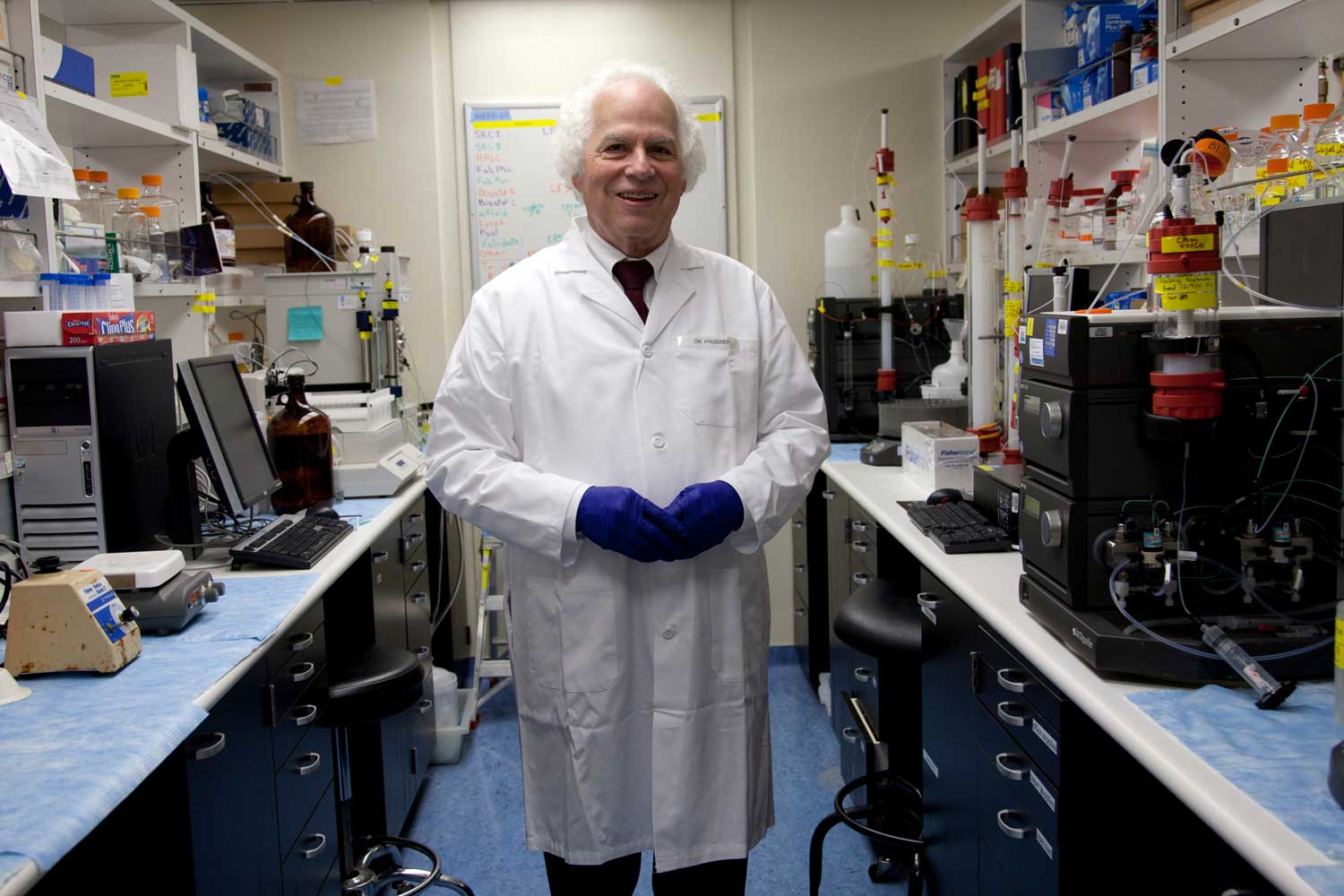 Stanely Prusiner wears a lab coat and stands in a lab.