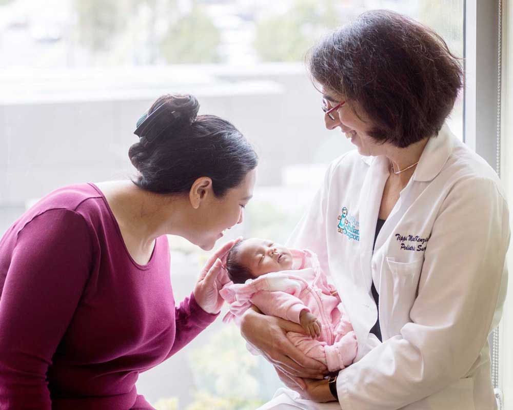Neonatal surgeon Tippi Mackenzie holds a newborn baby as her mother smiles and holds the baby's head.