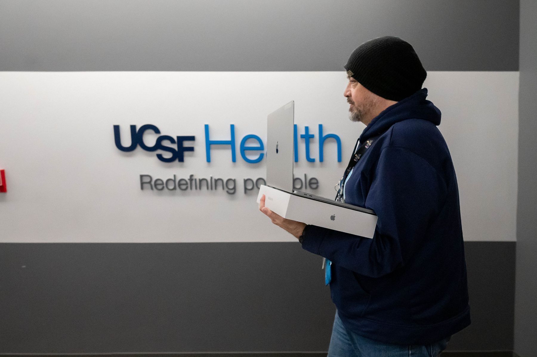 A man walking with a new laptop and box in hand, in front of a UCSF Health logo wall.