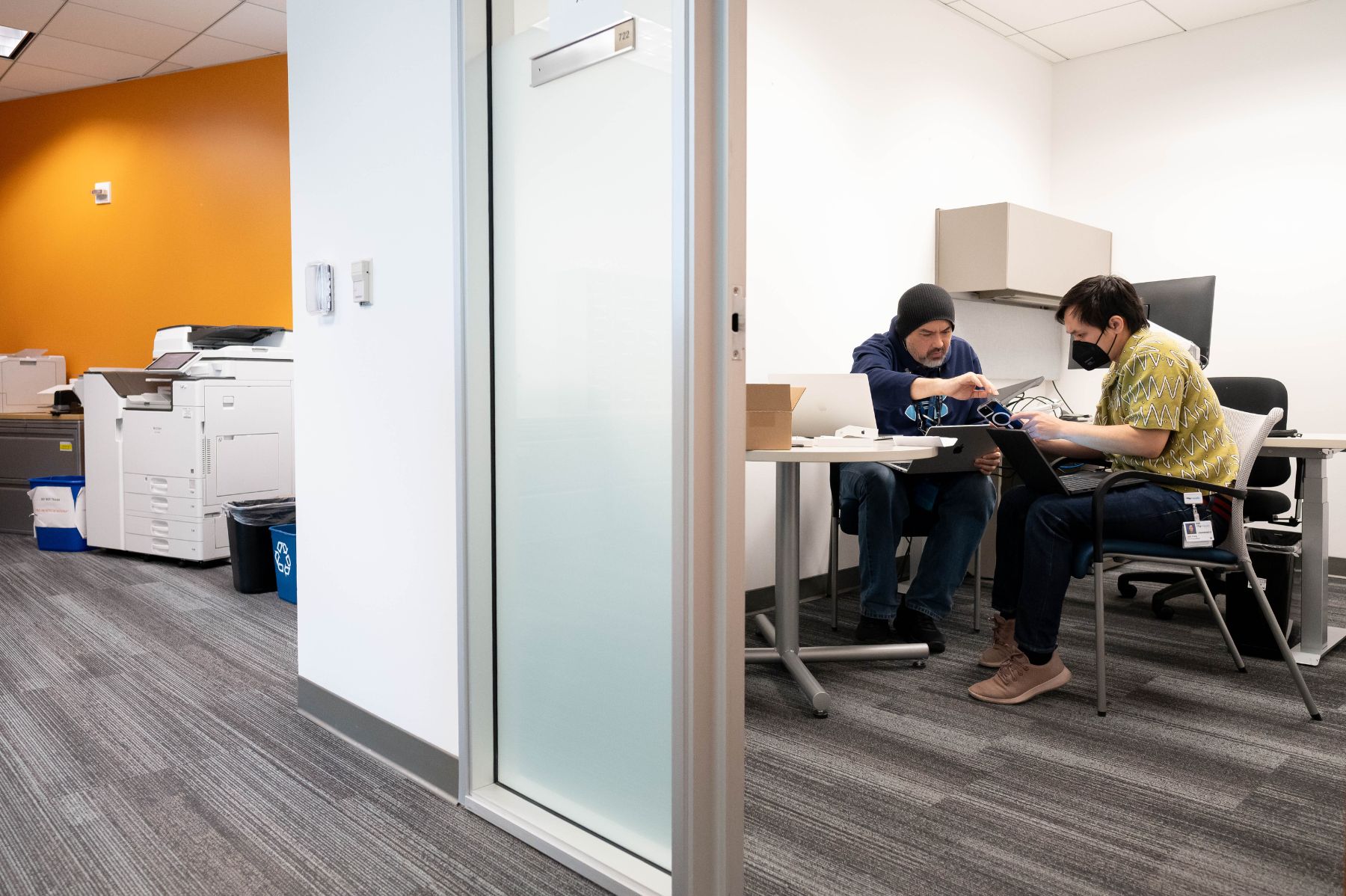 Two men working together on laptops at a small table in an office.