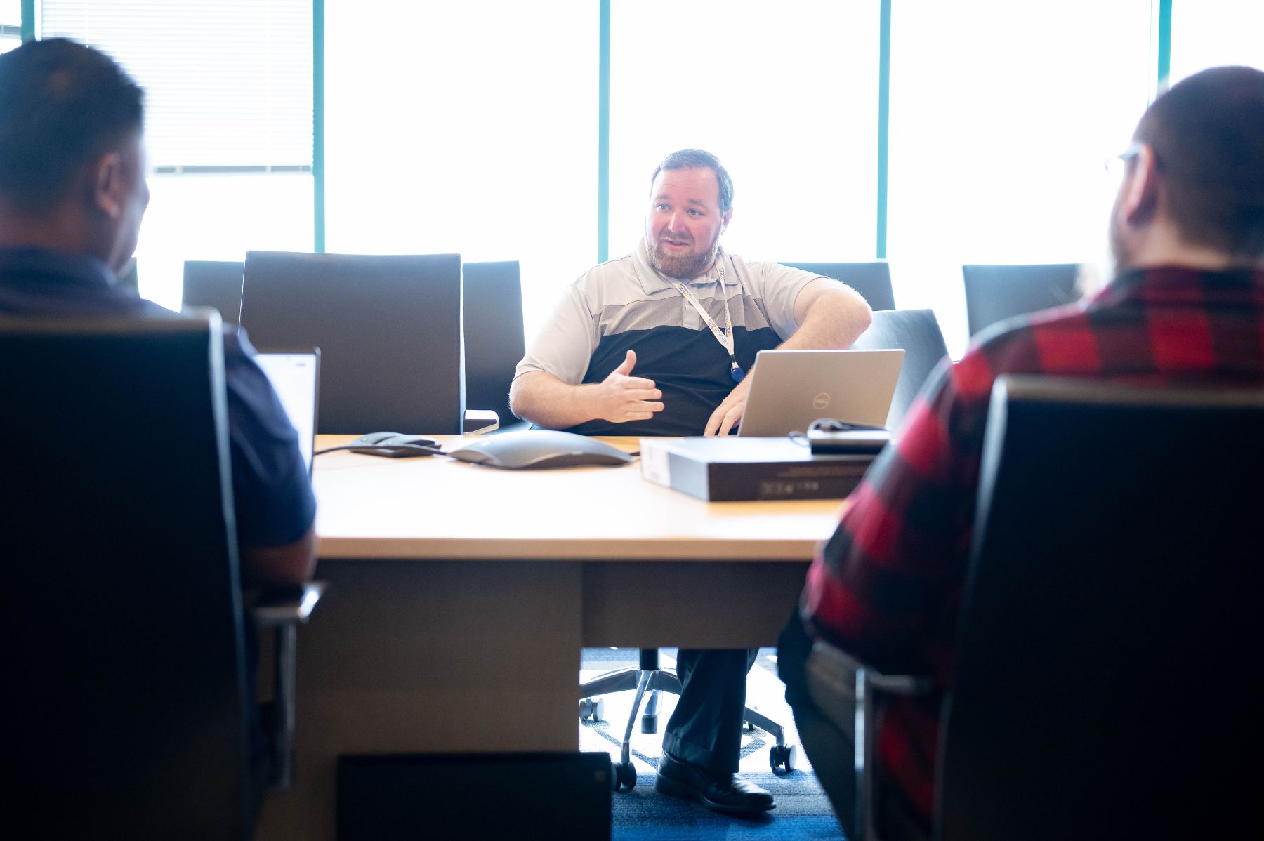 A man speaking and gesturing with his hands during a meeting, with two colleagues seen from behind.