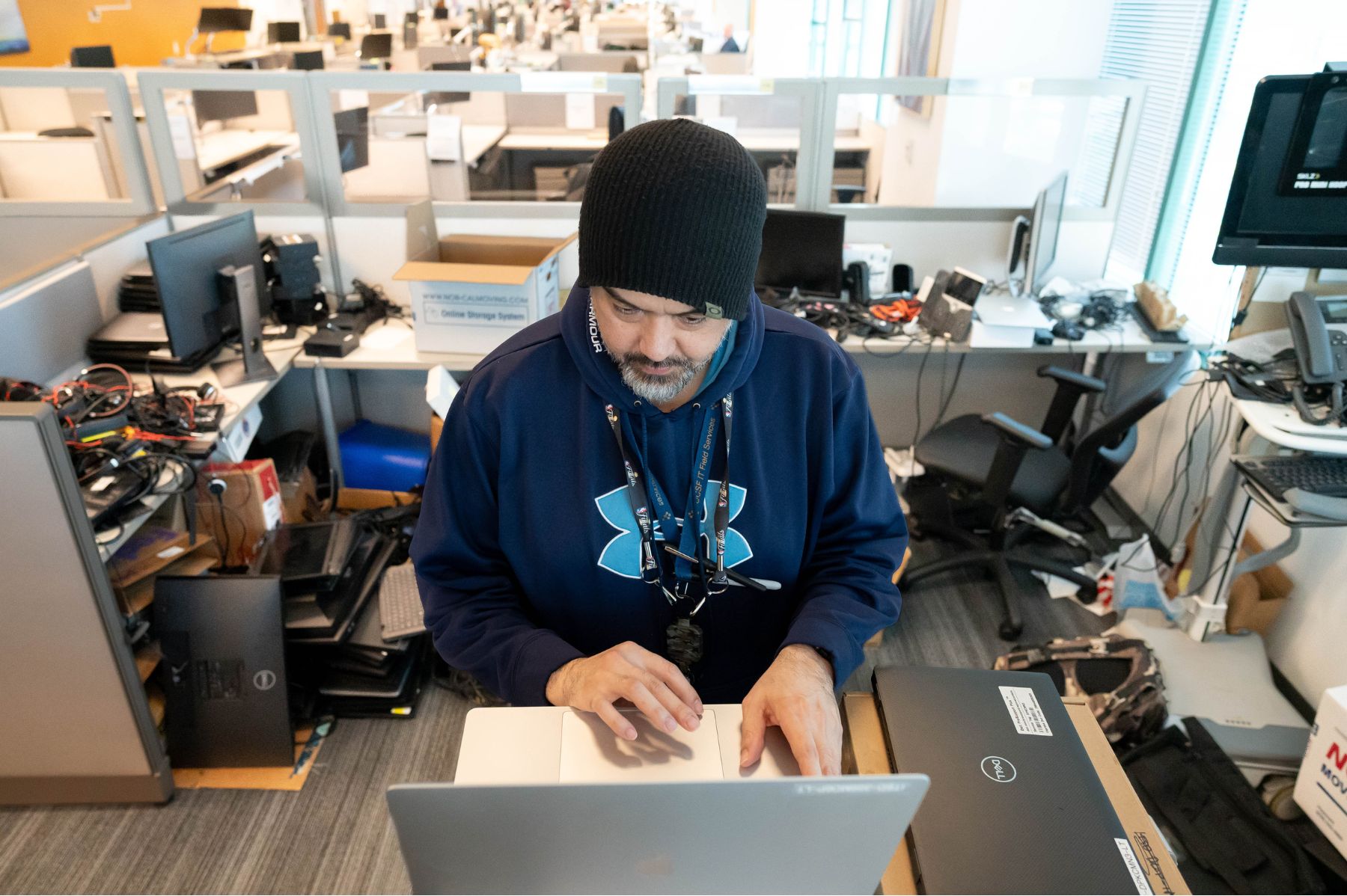 A man with a beanie sitting in a cluttered office cubicle, working on a laptop.