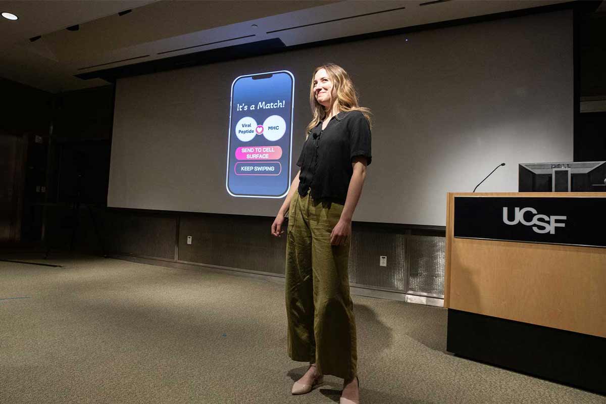 2025 Grad Slam winner Sophia Miliotis stands onstage in front of a projection screen that shows a graphic representation of the dating app Tinder, where cells are either chosen to be sent to the surface, or "swiped on".