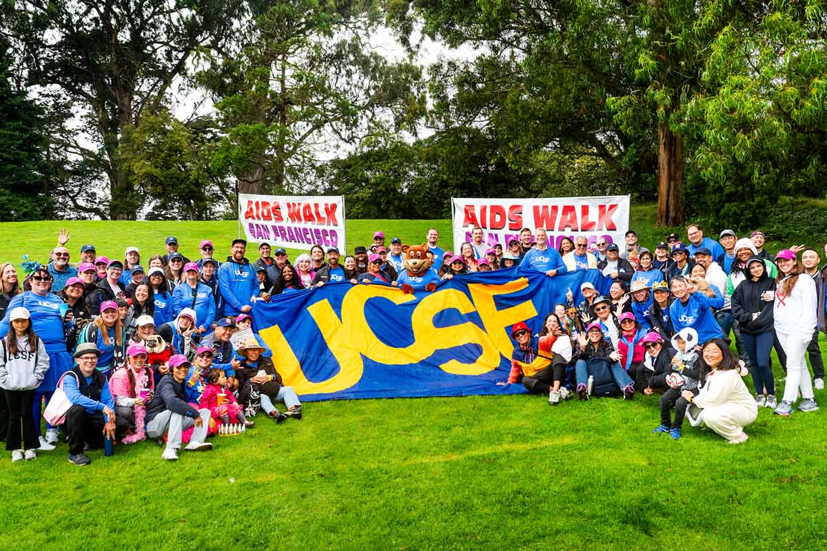 A large group of people at the AIDS walk event hold a large sign that reads "U C S F" and two large banners that read "AIDS Walk."